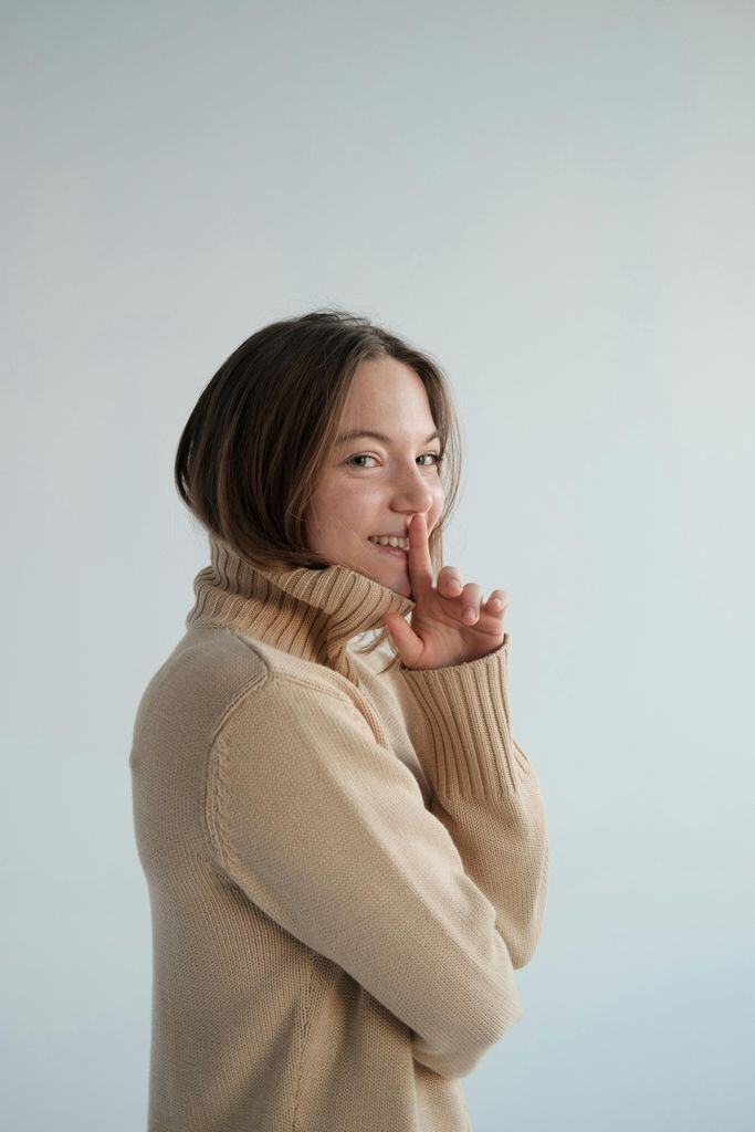 Side view of positive female wearing beige turtleneck covering mouth with finger against white background and looking at camera